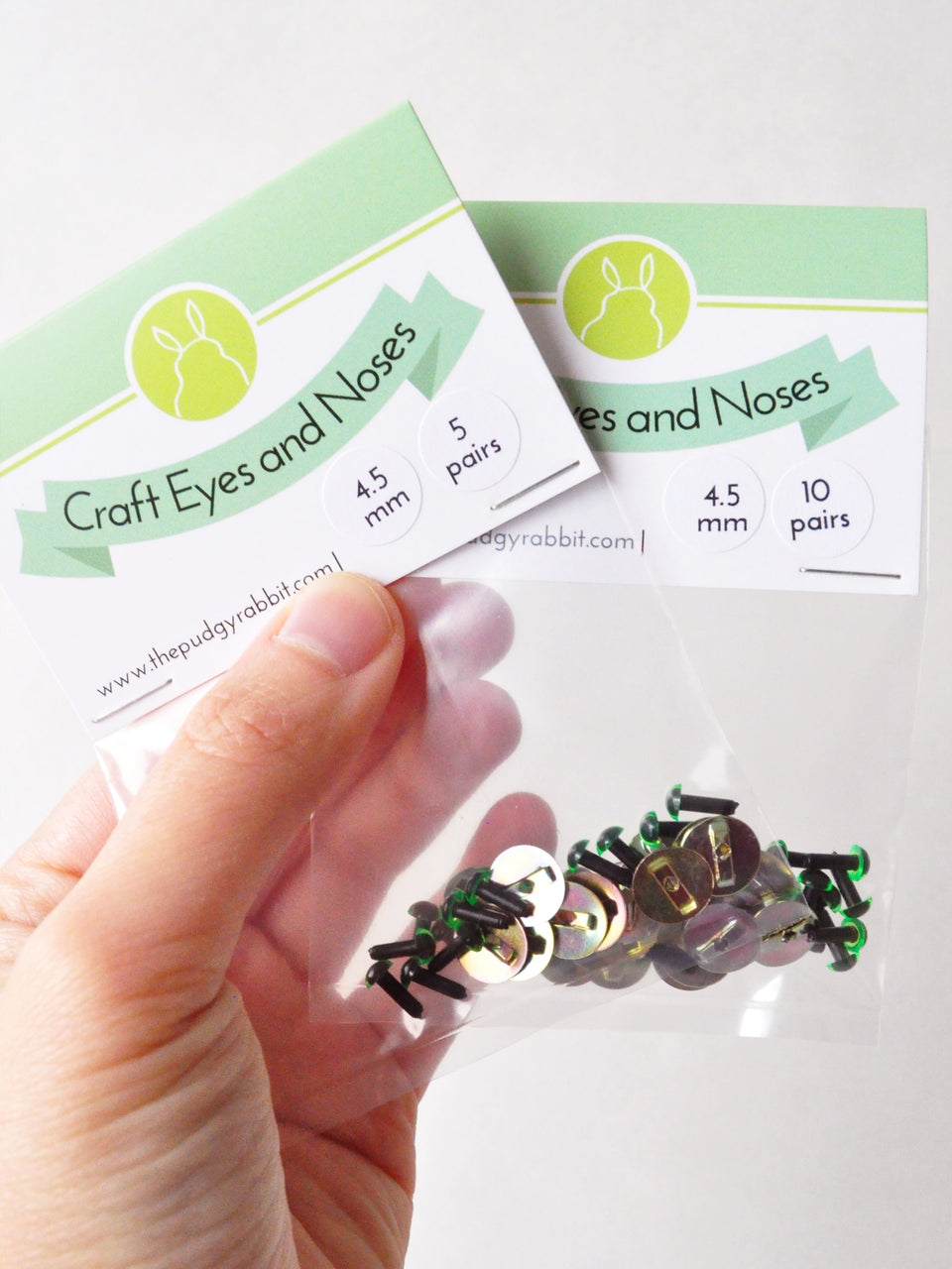 Packaged green craft eyes with a hand holding the packaging against a white background.