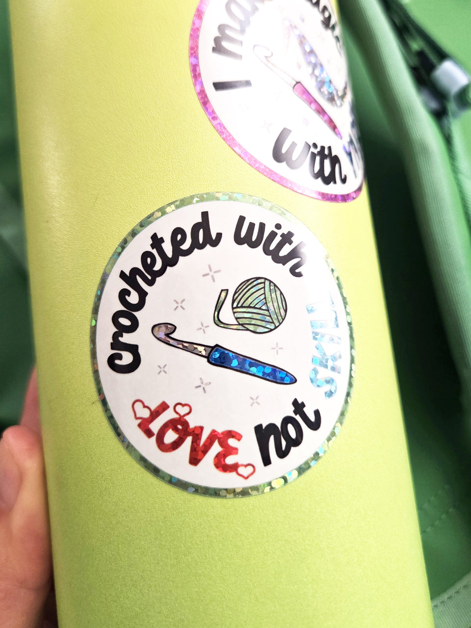 Glittery circle sticker that says "Crocheted with love, not skill" with a picture of a crochet hook and yarn ball in the center.