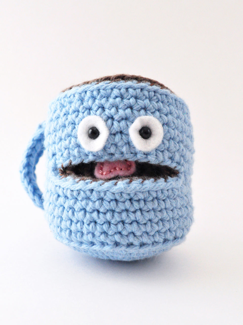 Blue crochet coffee mug amigurumi with googly eyes and a pink tongue on a white background, pattern by The Pudgy Rabbit