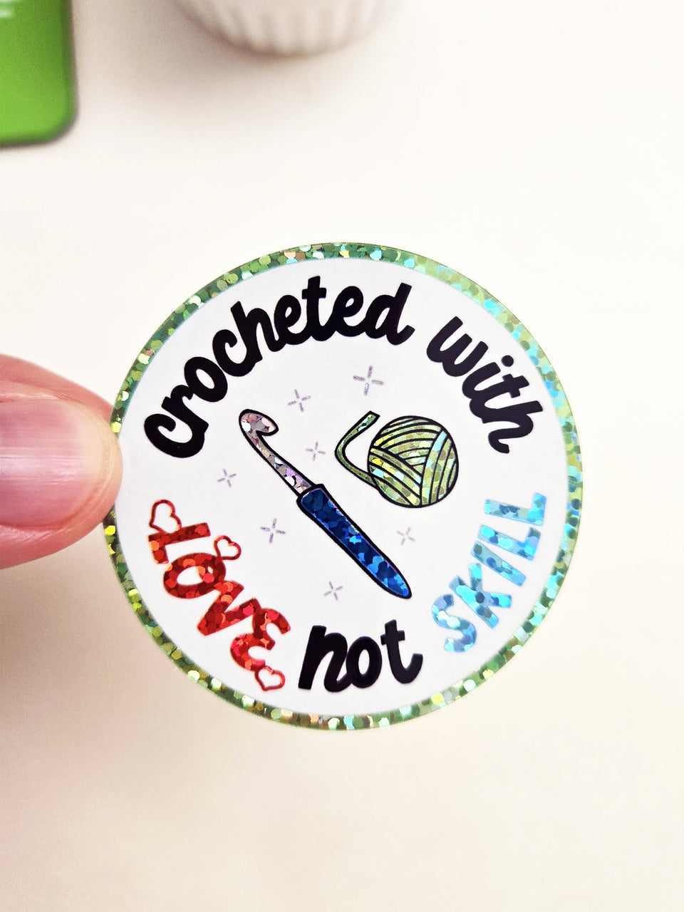 Round sticker with glitter accents that says "Crocheted with love, not skill" with a picture of a crochet hook and yarn ball in the center.