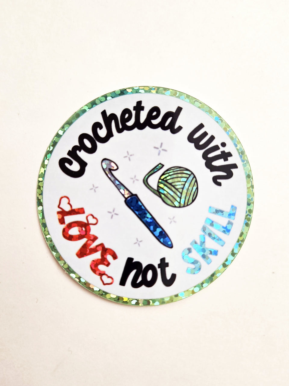Round glittery sticker with the saying, "Crocheted with love, not skill" and a  crochet hook and yarn ball in the center.