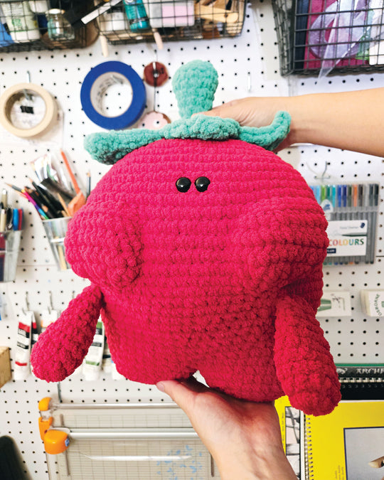 Crochet strawberry plush toy held in front of a wall with stationery items.