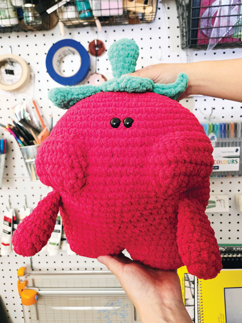 Hand holding a crochet strawberry plush toy with green leaves against a pegboard background.