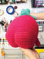 Hand holding a crochet strawberry butt in front of a pegboard