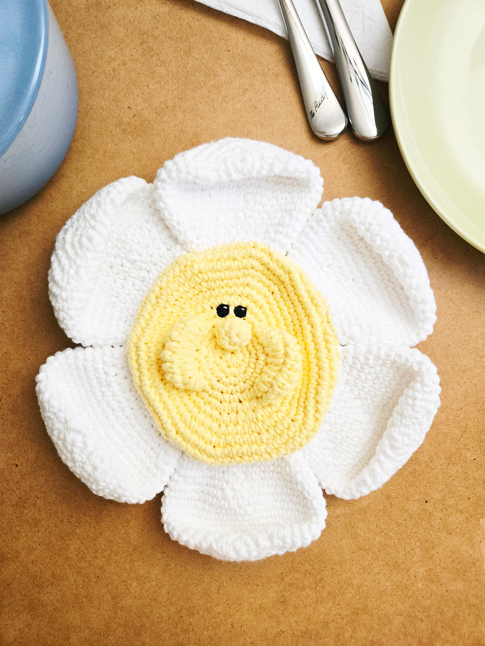 Crocheted flower trivet with a yellow center and black eyes on a brown surface.