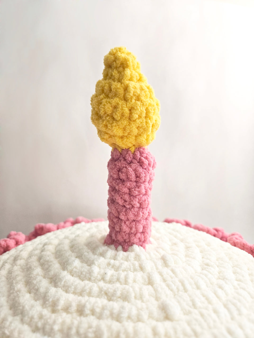Close-up of a crocheted pink candle with yellow flame on top of a white cake amigurumi 