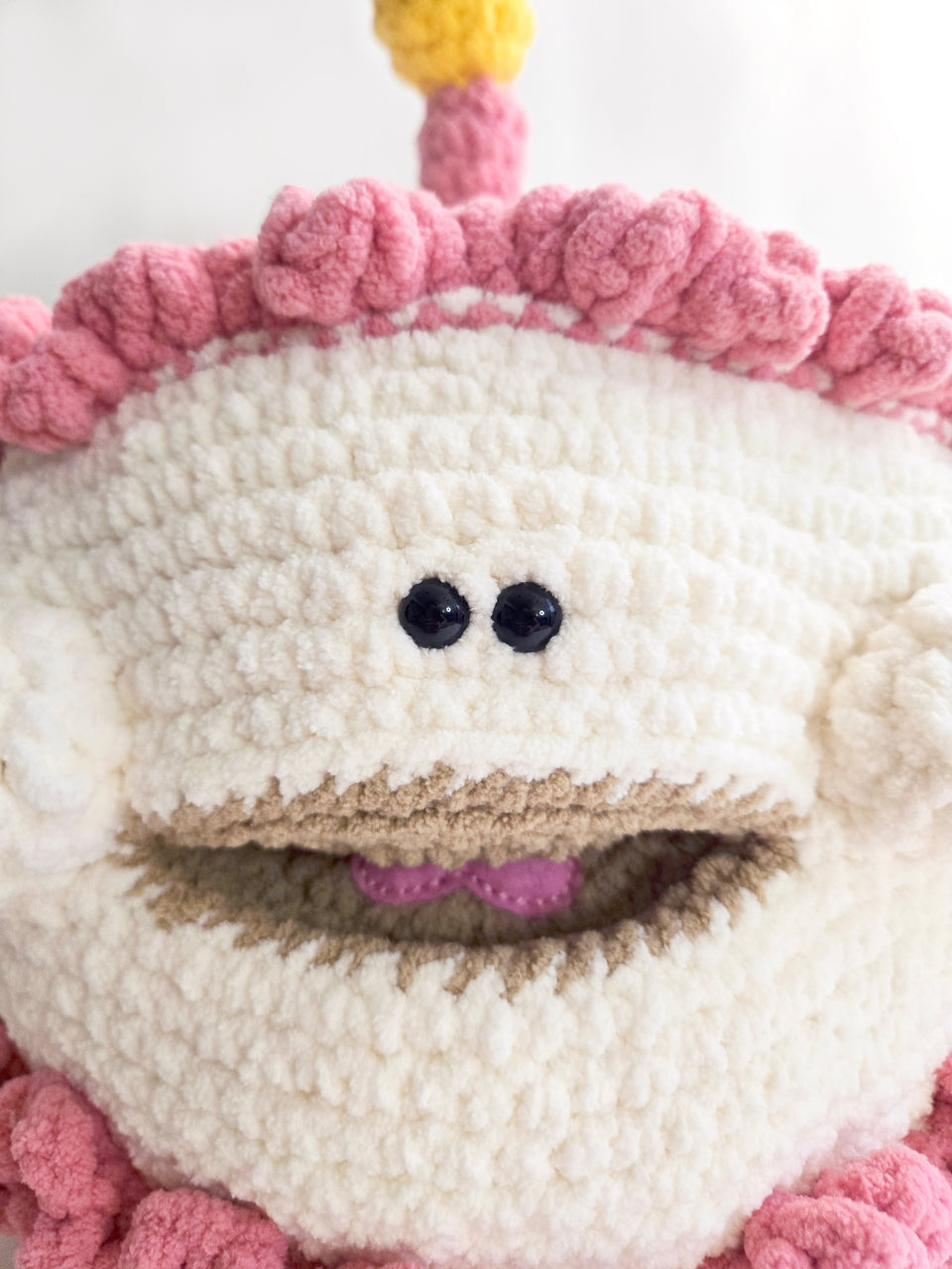 Close-up of a crocheted birthday cake with pink frosting, chubby cheeks, and open smile.