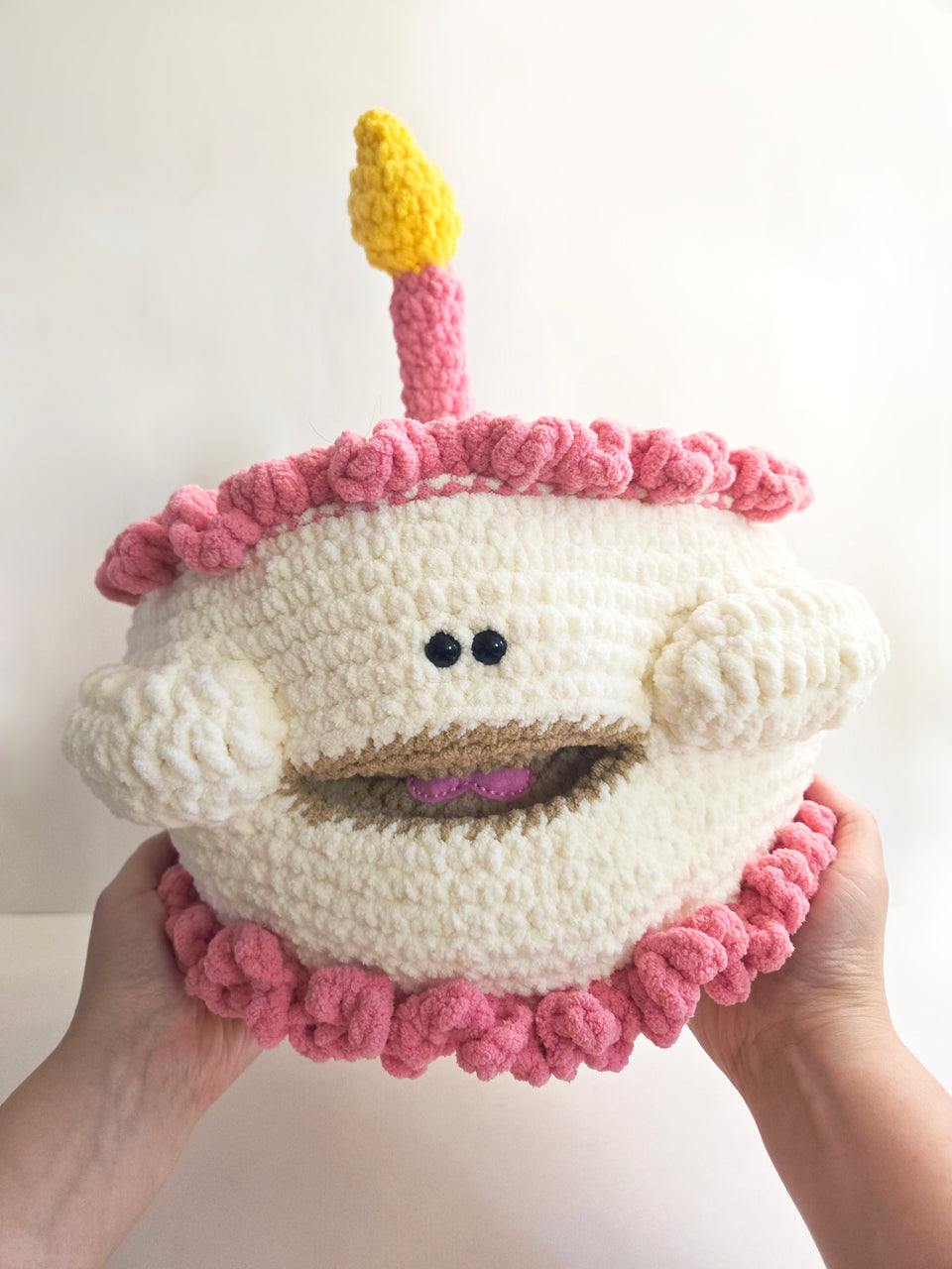 Crocheted birthday cake  plushie with a candle held by hands against a white background