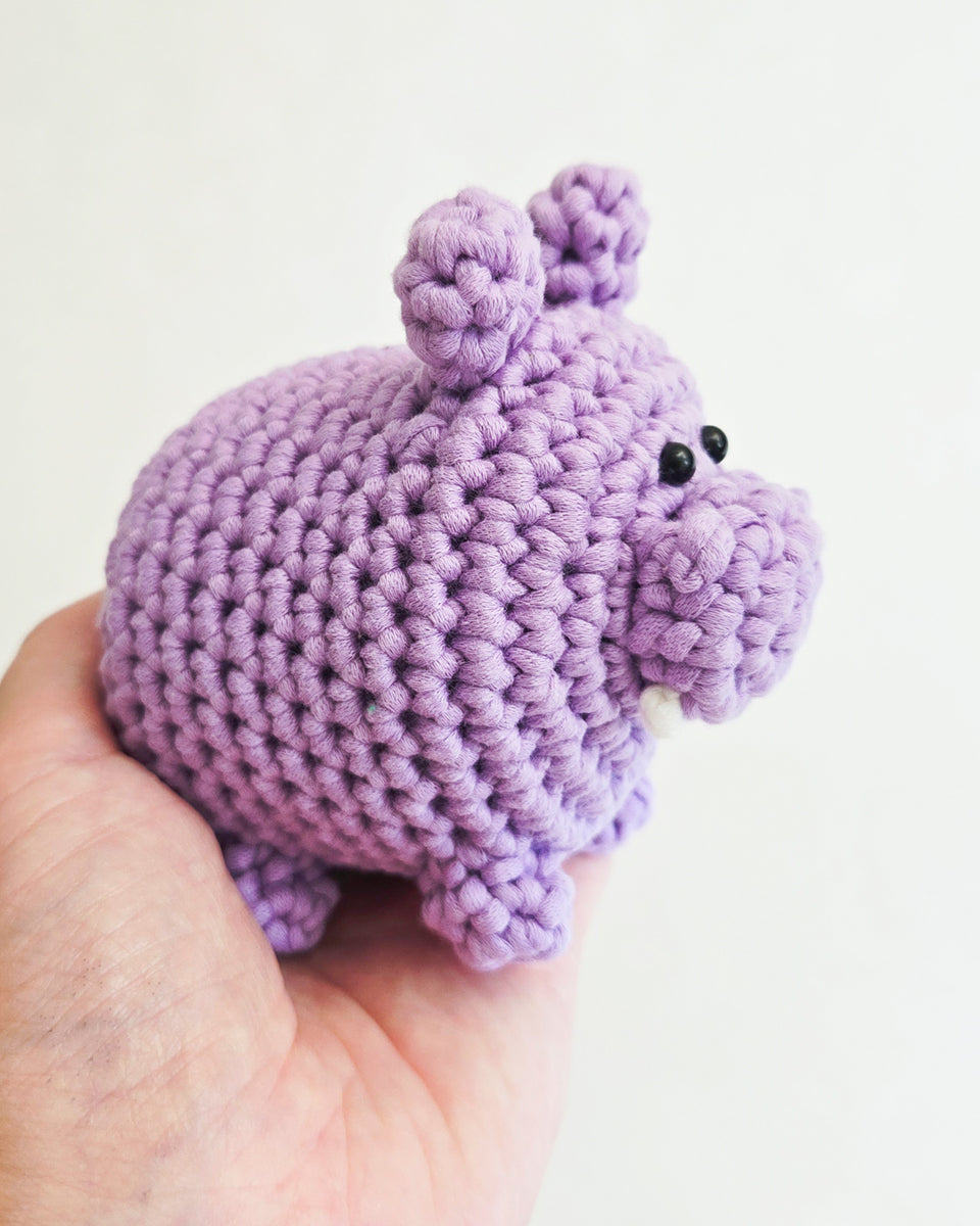 A small crocheted hippo toy in light purple yarn, held in a person's hand against a plain light background.