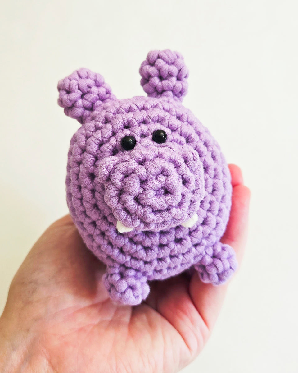 A small crocheted hippo toy made from a crochet kit, is being held in a person's hand against a plain light background. The hippo is made from lavender-purple yarn and has black bead eyes and small white yarn teeth.