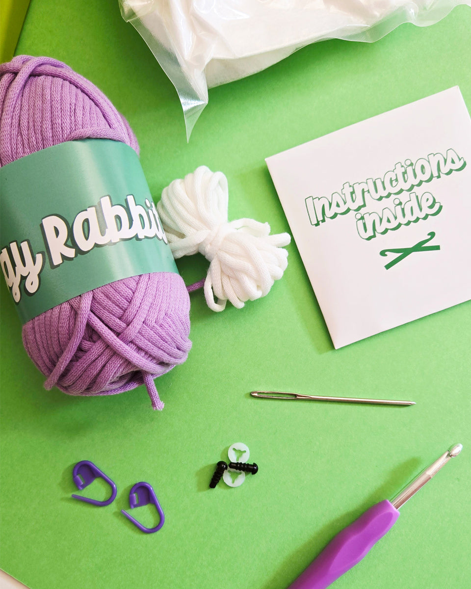 A flat-lay of crochet kit components on a bright green background, including a purple ball of yarn with a teal label reading 'The Pudgy Rabbit', white yarn, a crochet hook with a purple handle, purple stitch markers, a metal yarn needle, and a white packet that says 'Instructions inside'.