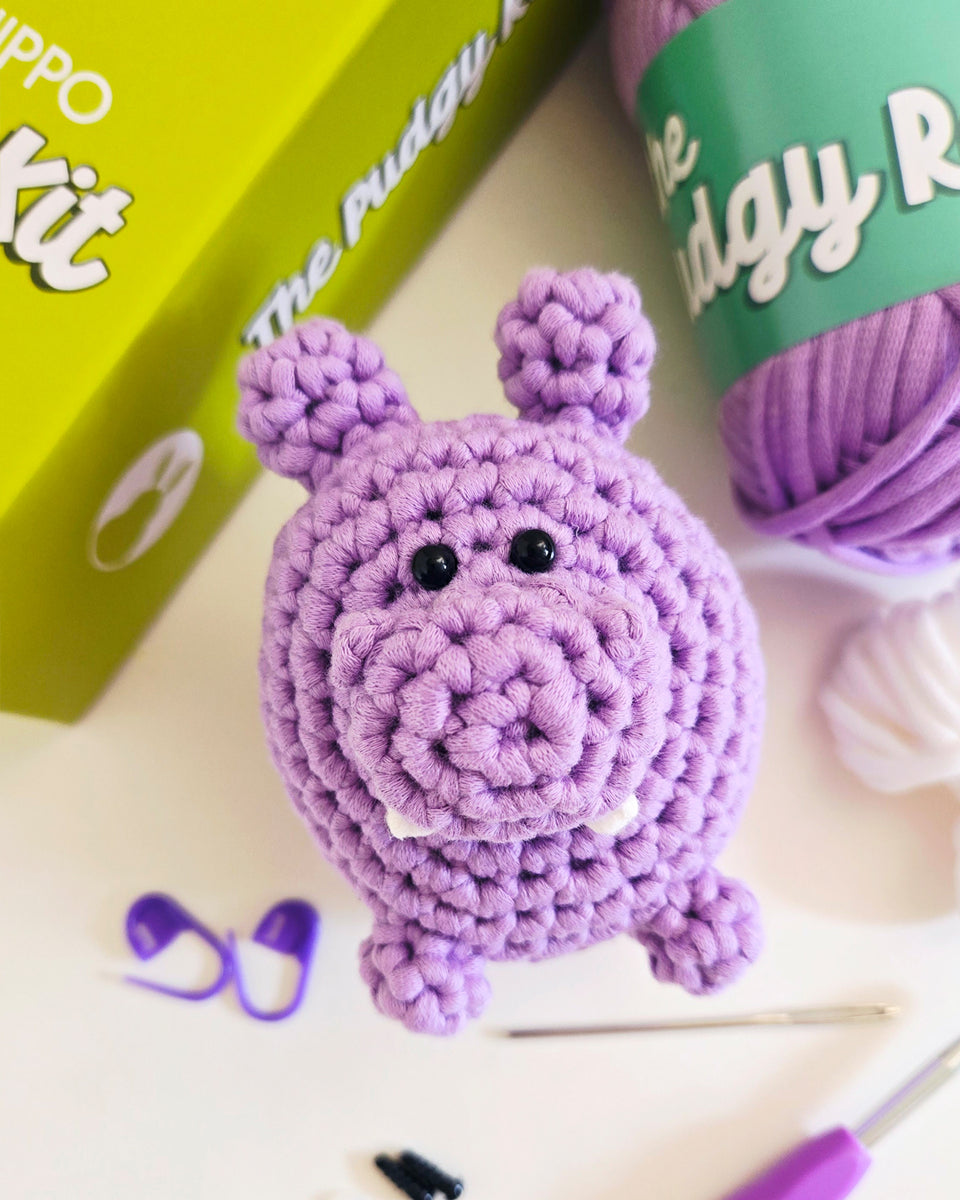 A crocheted purple hippo toy sits on a white surface with crochet hooks, yarn, and a green box partially visible in the background. The box reads 'The Pudgy Rabbit crochet kit'.