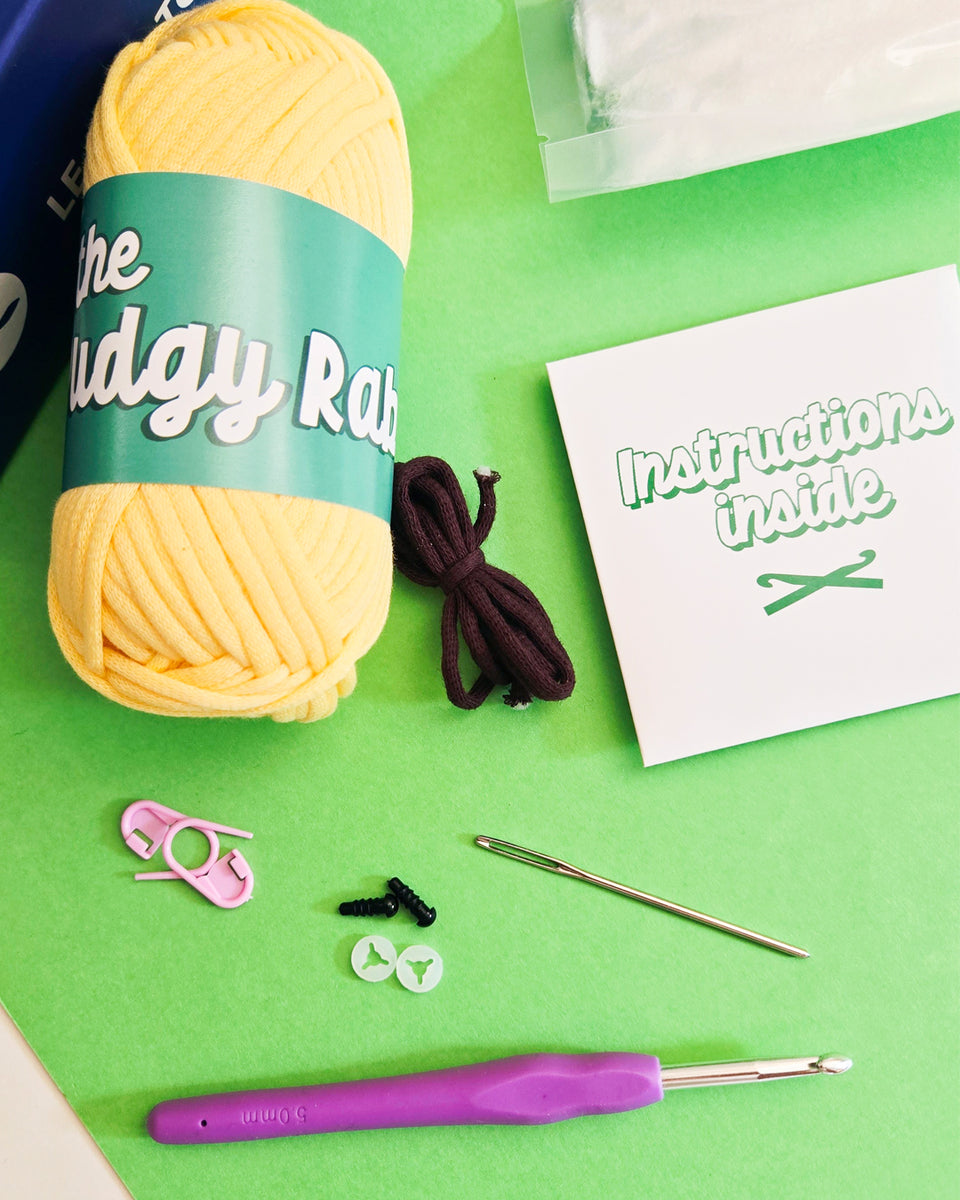 A dog crochet kit by The Pudgy Rabbit laid out on a bright green background, including a large skein of yellow yarn with a teal label, a purple-handled crochet hook, pink stitch markers, safety eyes, a metal yarn needle, and a white packet that reads 'Instructions inside'.
