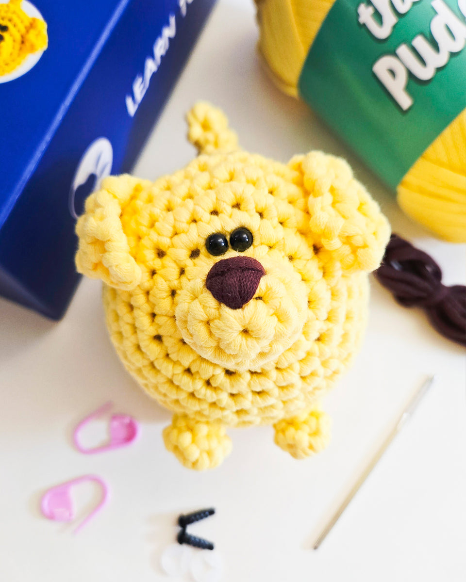 A crocheted yellow puppy with black bead eyes and a dark brown nose, displayed on a white surface with knitting accessories and yarn balls around it. Dog made from crochet kit by The Pudgy Rabbit.