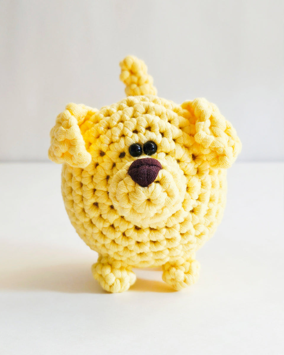 A yellow crocheted dog toy floppy ears, black bead eyes, and a dark brown nose, standing on a light gray background. Dog made from learn to crochet kit by The Pudgy Rabbit.