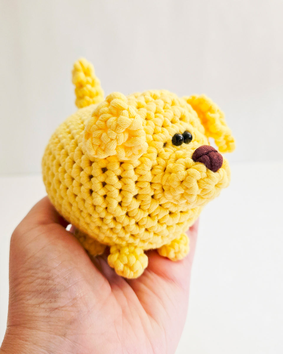 A small yellow crocheted plush dog, made from a crochet kit, being held in a person's hand against a plain light background.