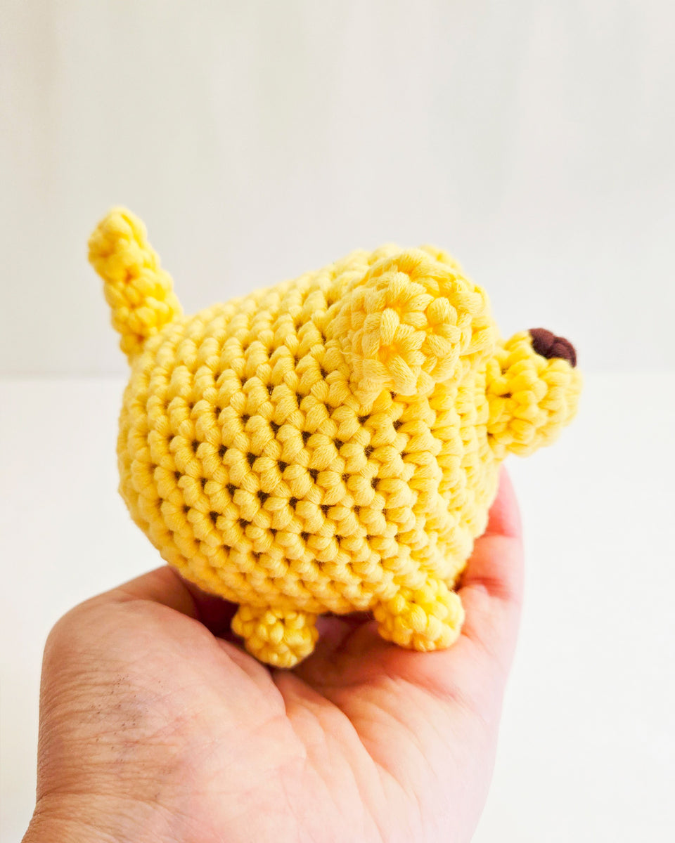 Side view of a small yellow crocheted dog toy being held in a person's hand against a plain light background.