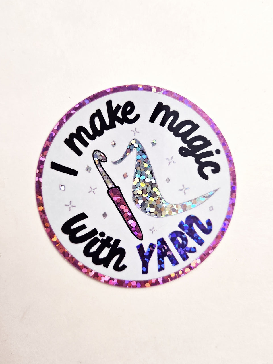 Circle shaped sticker with glitter accents that says "I make magic with yarn" with a crochet hook in the middle.
