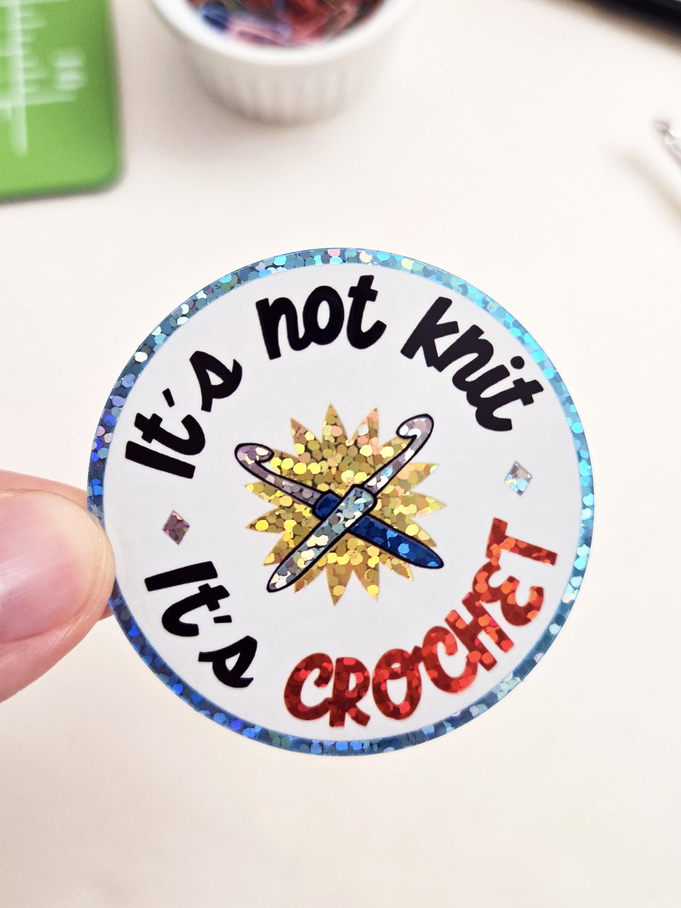 A round sticker held by a finger, featuring the text 'It's not knit, it's crochet' in black and red glitter-style lettering around a central illustration of a blue and gold crochet hook.