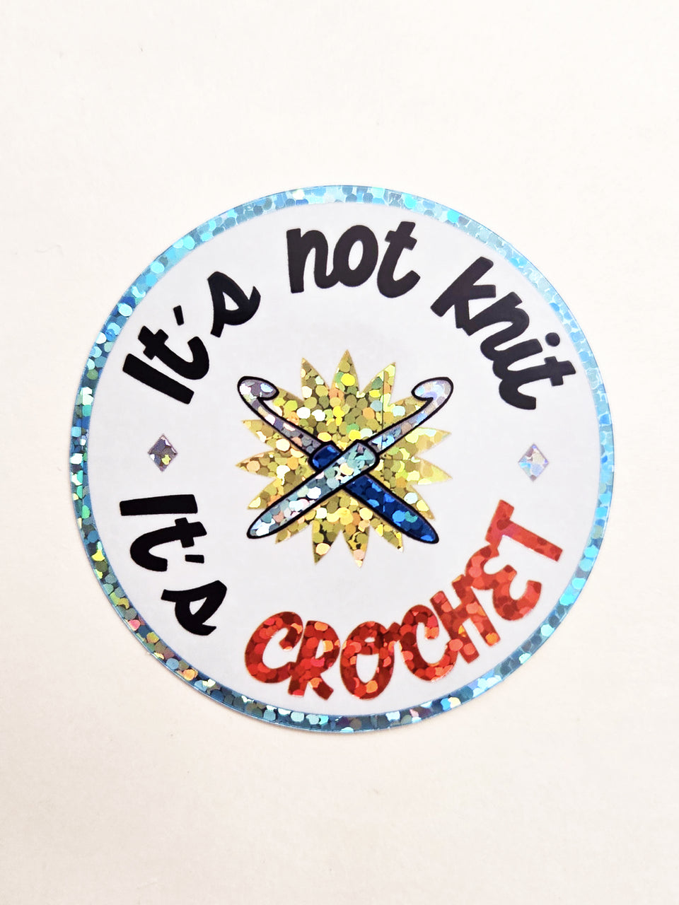 A round sticker with a holographic glitter border and the text 'It's not KNIT it's CROCHET' in black and red lettering, featuring a blue crochet hook graphic in the center on a white background.