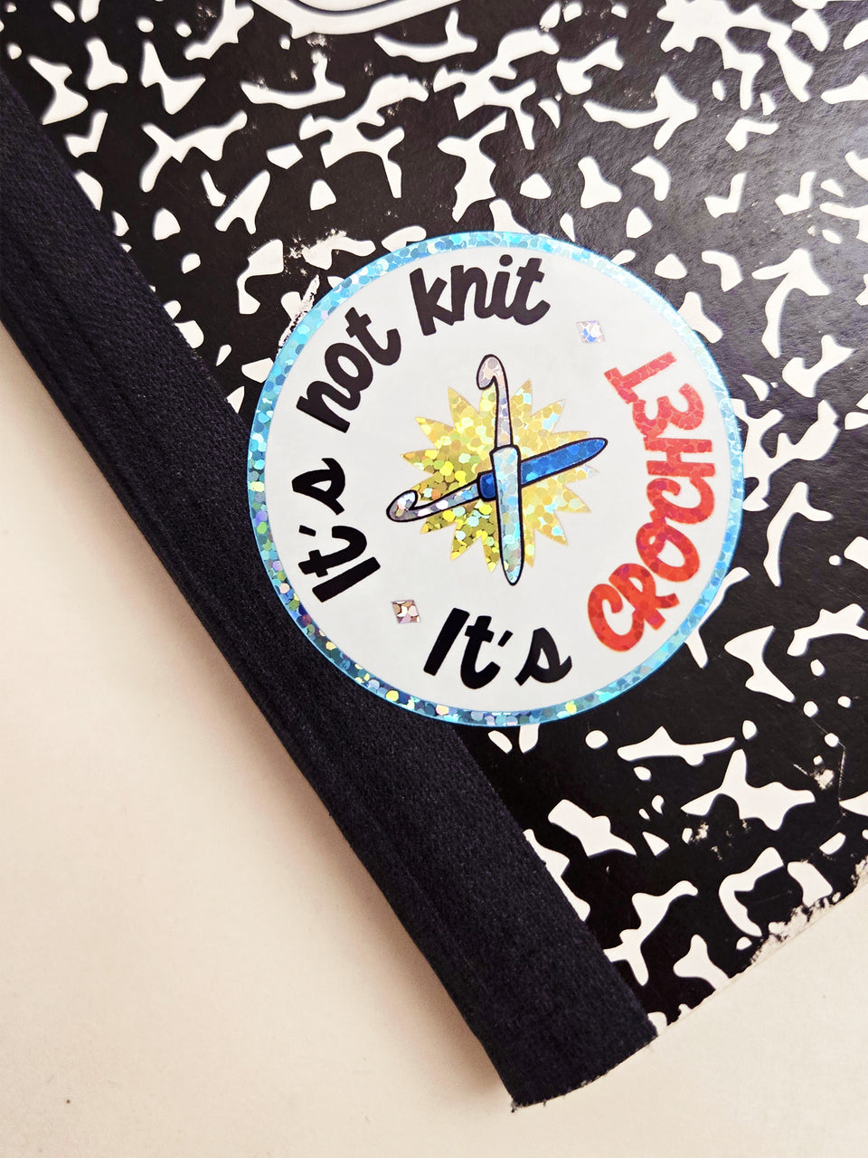 A close-up of a round sticker placed on a black-and-white patterned composition notebook. The sticker reads “It’s not knit It’s crochet” with a crochet hook illustration in the center.