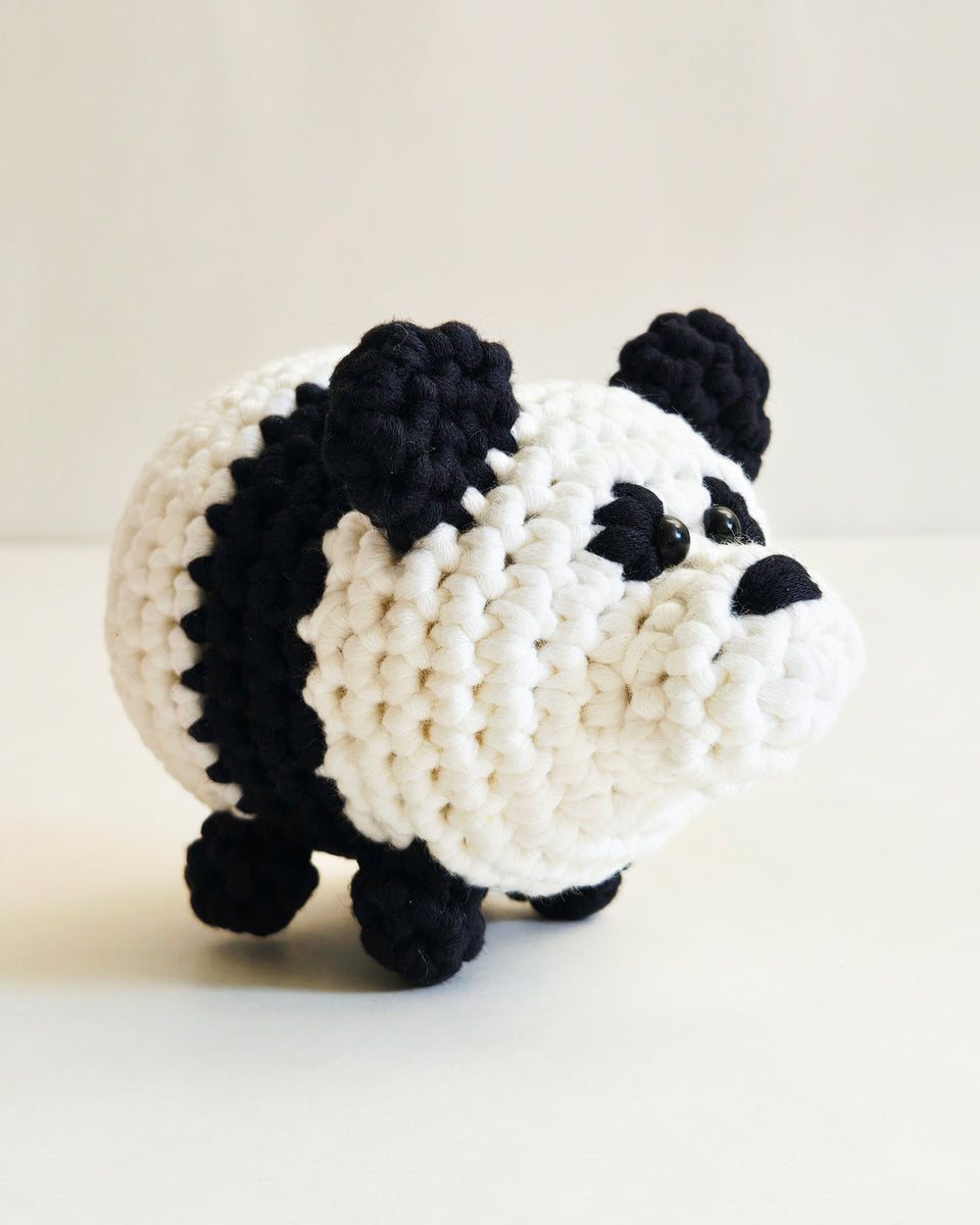 A handmade crocheted panda amigurumi photographed on a light beige background. Panda made from crochet kit for beginners by The Pudgy Rabbit.