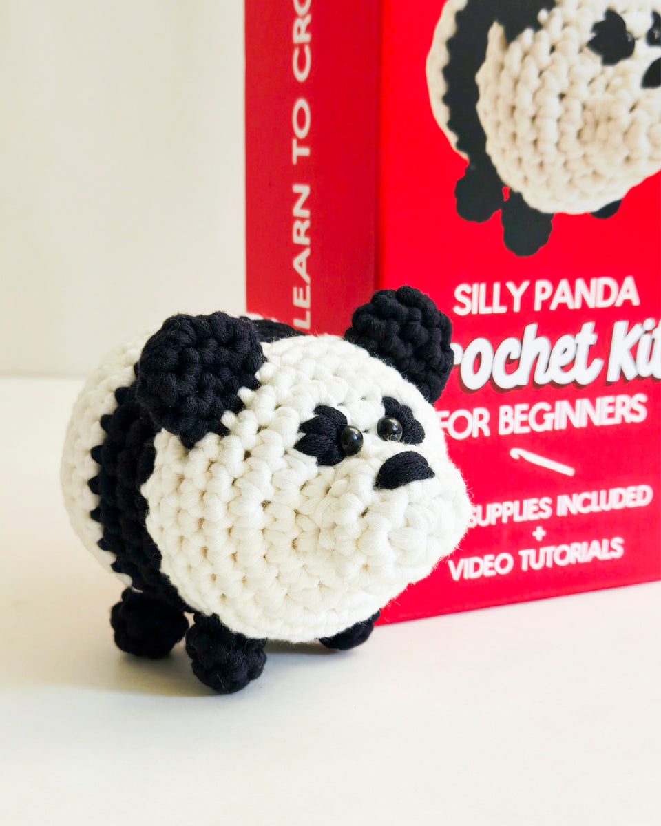 A crocheted panda toy in black and white sits in front of a red crochet kit titled 'Silly Panda Crochet Kit for Beginners'.