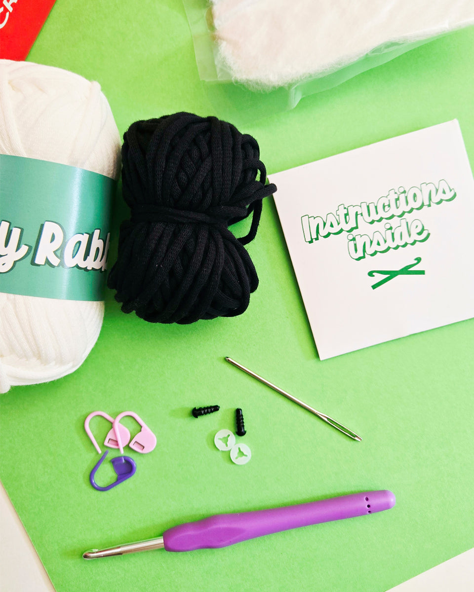 A flat-lay of crochet starter supplies on a bright green background, including a ball of white yarn, a ball of black yarn, a purple-handled crochet hook, pink and purple stitch markers, black thread, a metal needle, and a small card that reads 'Instructions inside'. Supplies from a crochet kits by The Pudgy Rabbit.