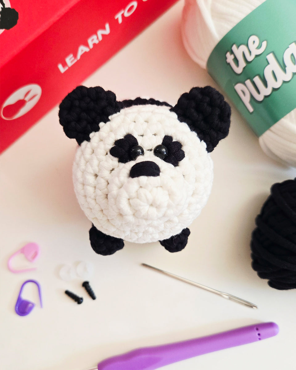 A crocheted panda from a diy crochet kit sits on a white surface with crochet tools and yarn around it. The panda is white with black ears, eyes, and paws.