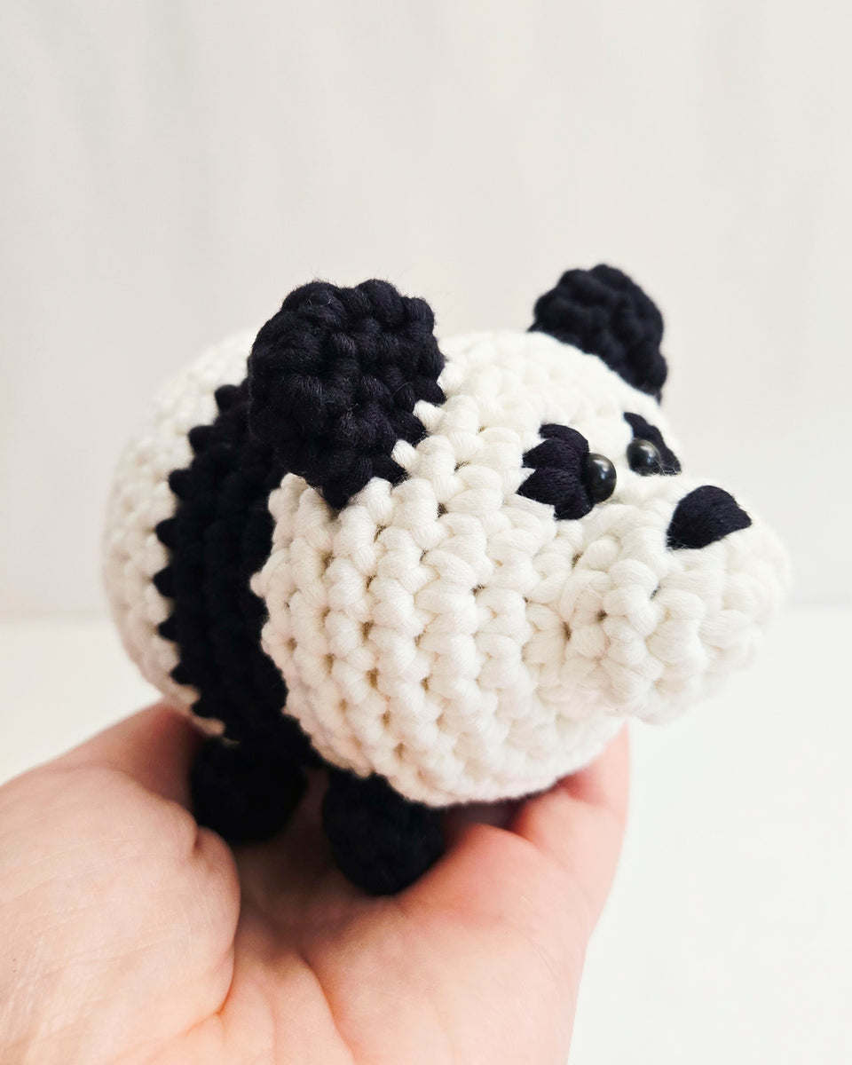 A small crocheted panda toy being held in a person's hand against a soft white background. Made from a DIY crochet kit by The Pudgy Rabbit.