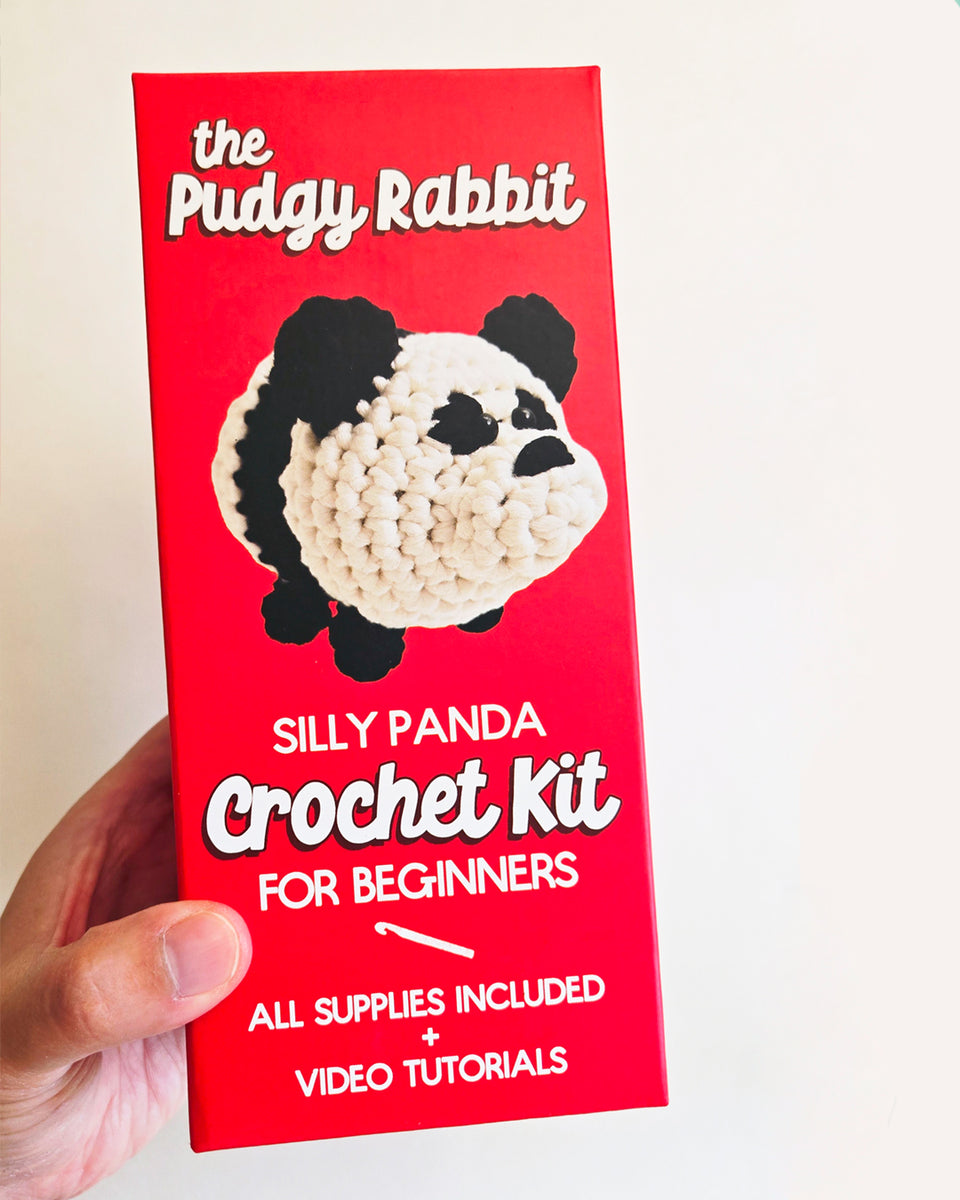 Hand holding a bright red box containing a panda crochet kit for beginners by The Pudgy Rabbit in front of a white background. 