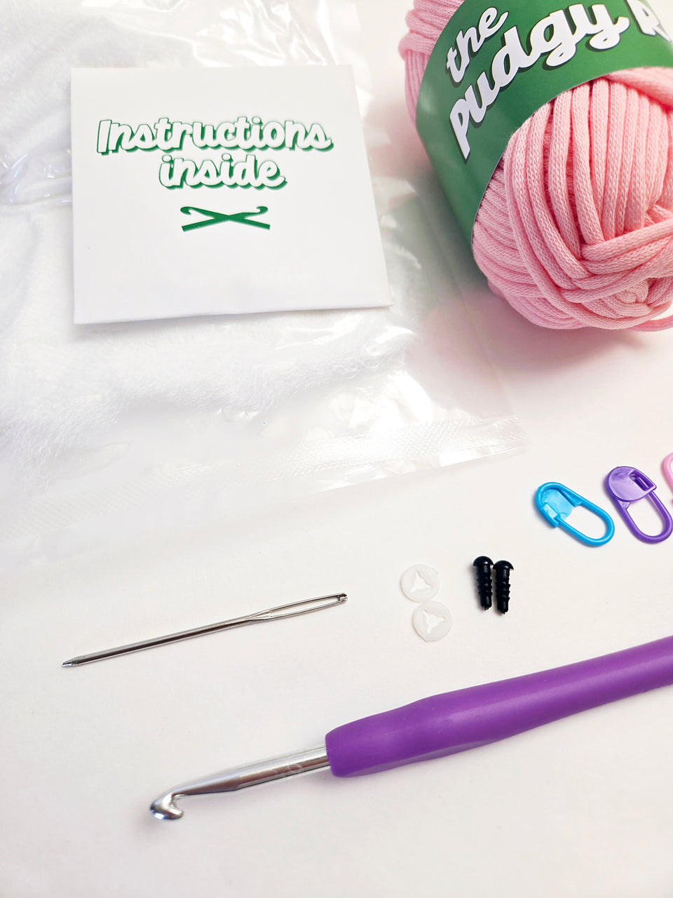 Close up of crochet kit supplies, including yarn, stuffing, crochet hook, yarn needle, safety eyes, stitch markers, and instruction packet.