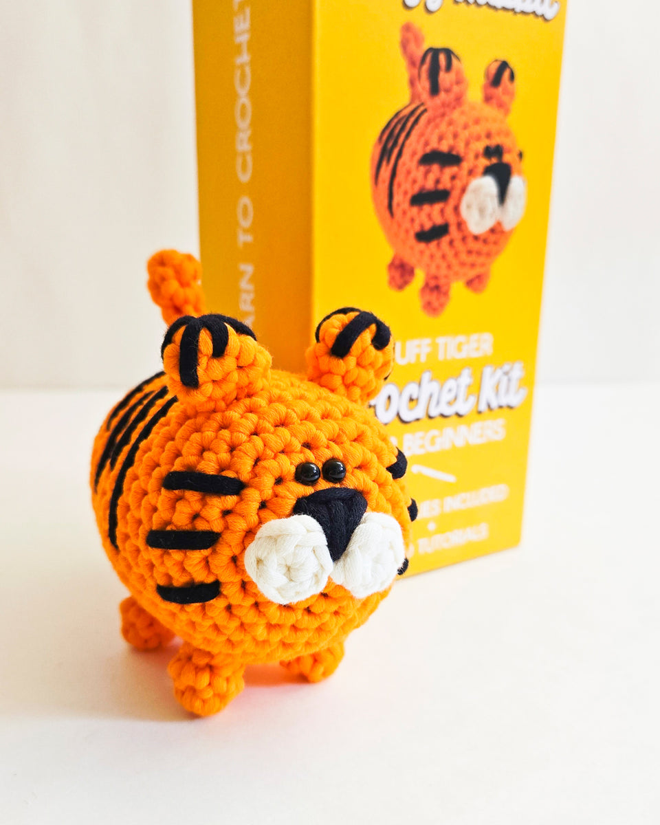 A crocheted orange tiger toy with black stripes sits in front of its bright yellow packaging box, which reads 'Learn to Crochet' and 'Tuff Tiger Crochet Kit'.