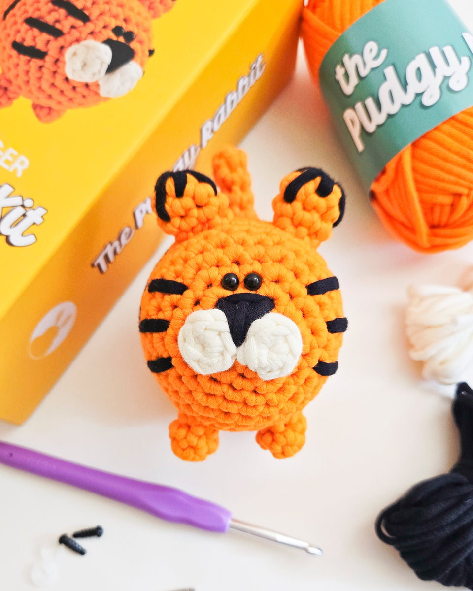 Crocheted orange tiger with black stripes, displayed with its yellow packaging box, an orange ball of yarn, a purple crochet hook, and other crafting materials on a white surface. Tiger from crochet kit by The Pudgy Rabbit.