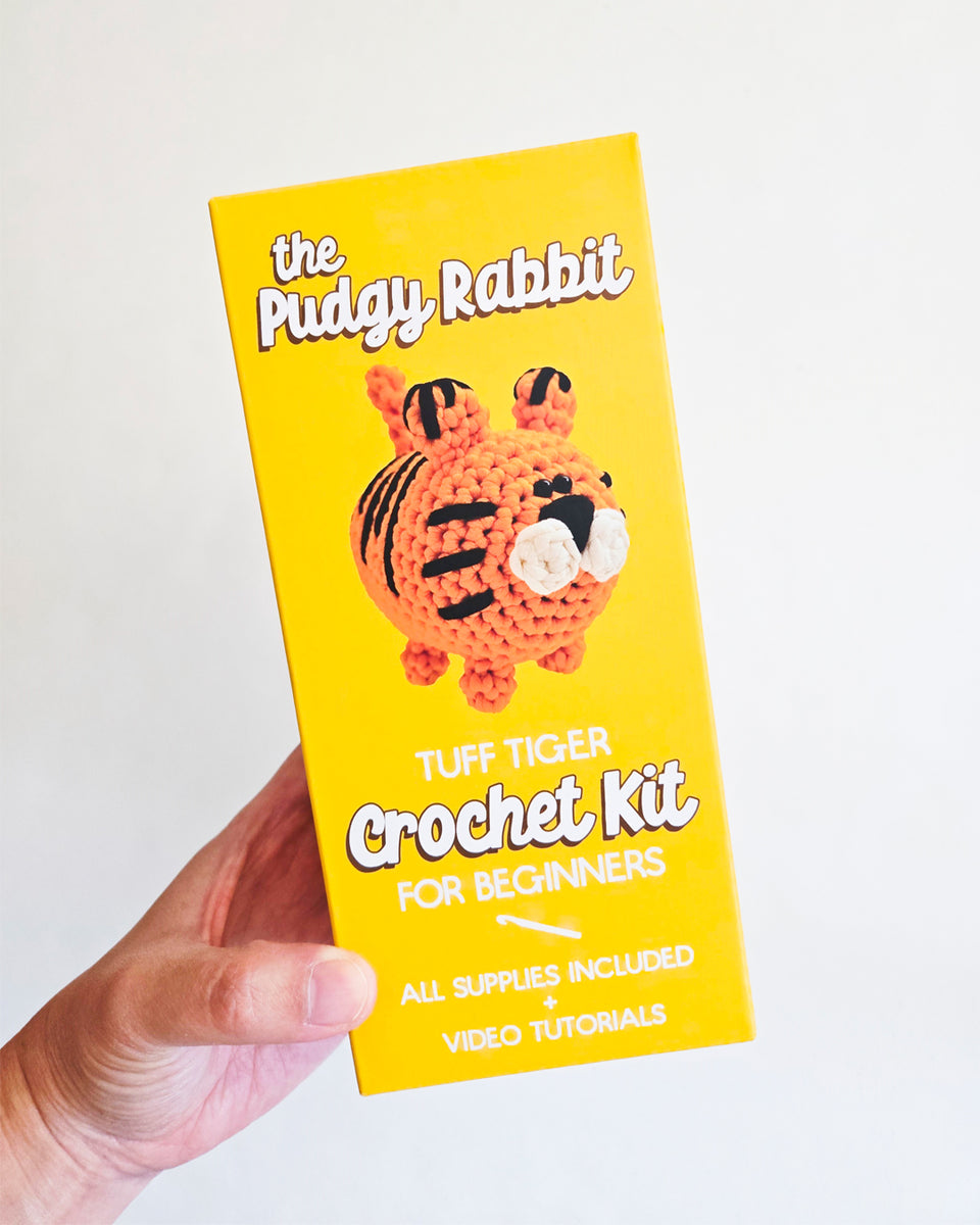 A hand holding a bright yellow box for 'the Pudgy Rabbit Tuff Tiger Crochet Kit for Beginners' against a plain white background.