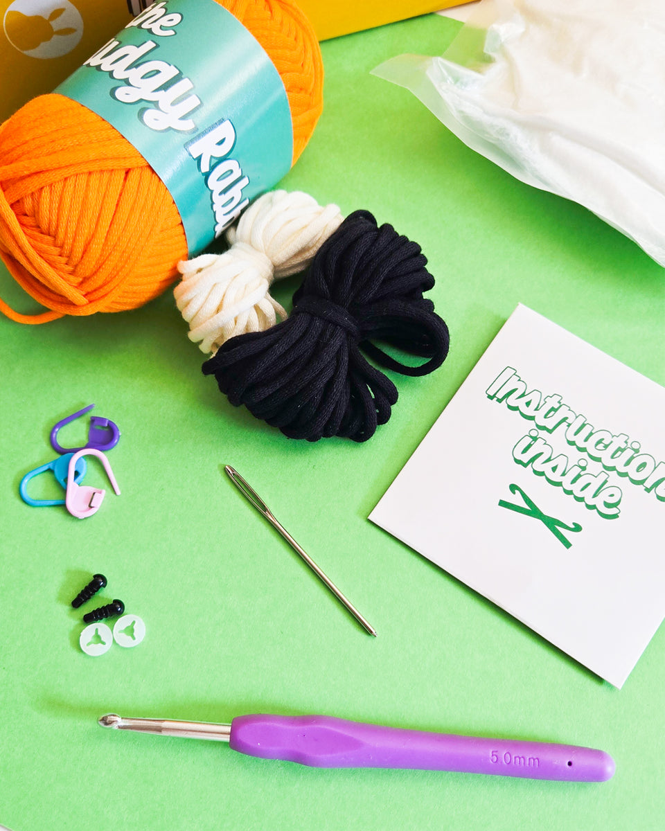 Learn to crochet kit laid out on a bright green background, including orange and cream yarn balls, black yarn, a purple-handled crochet hook, stitch markers, a needle, and a small instruction packet reading 'Instructions Inside.' 