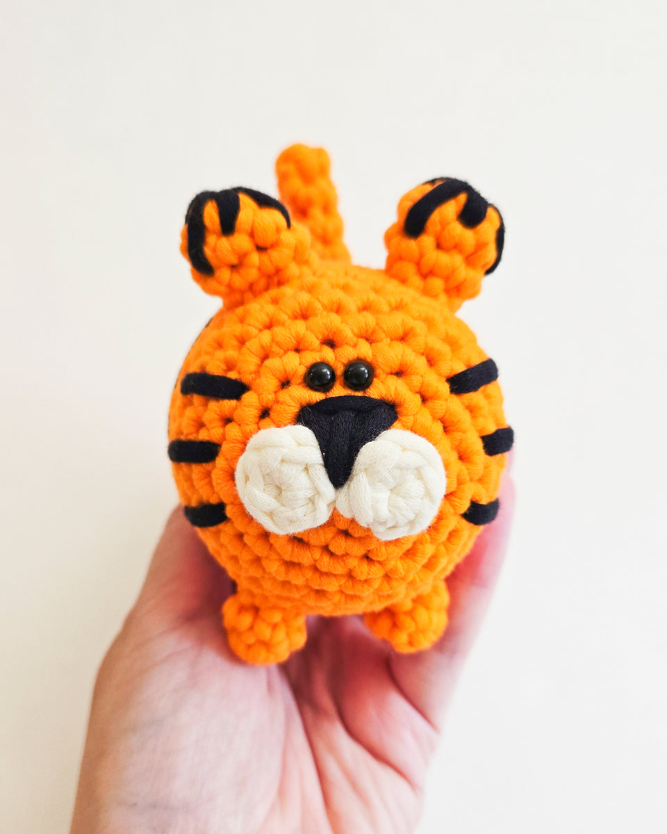 A small crocheted tiger toy being held in a person's hand against a plain light background. Featuring tiger made from beginner's crochet kit by The Pudgy Rabbit.