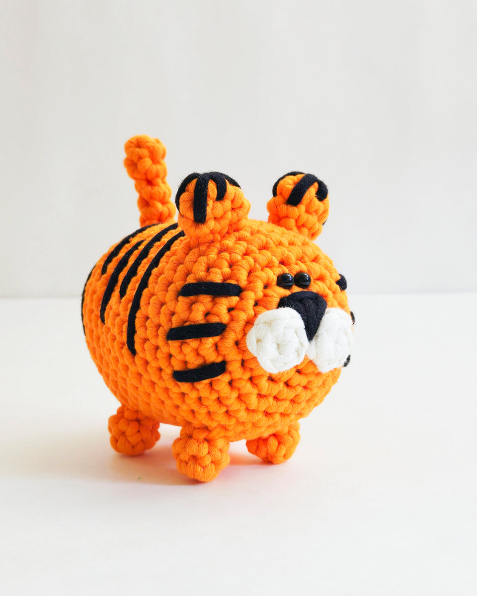 A crocheted orange tiger toy with black stripes, standing on a white surface against a white background. Tiger made from a learn to crochet kit by The Pudgy Rabbit.