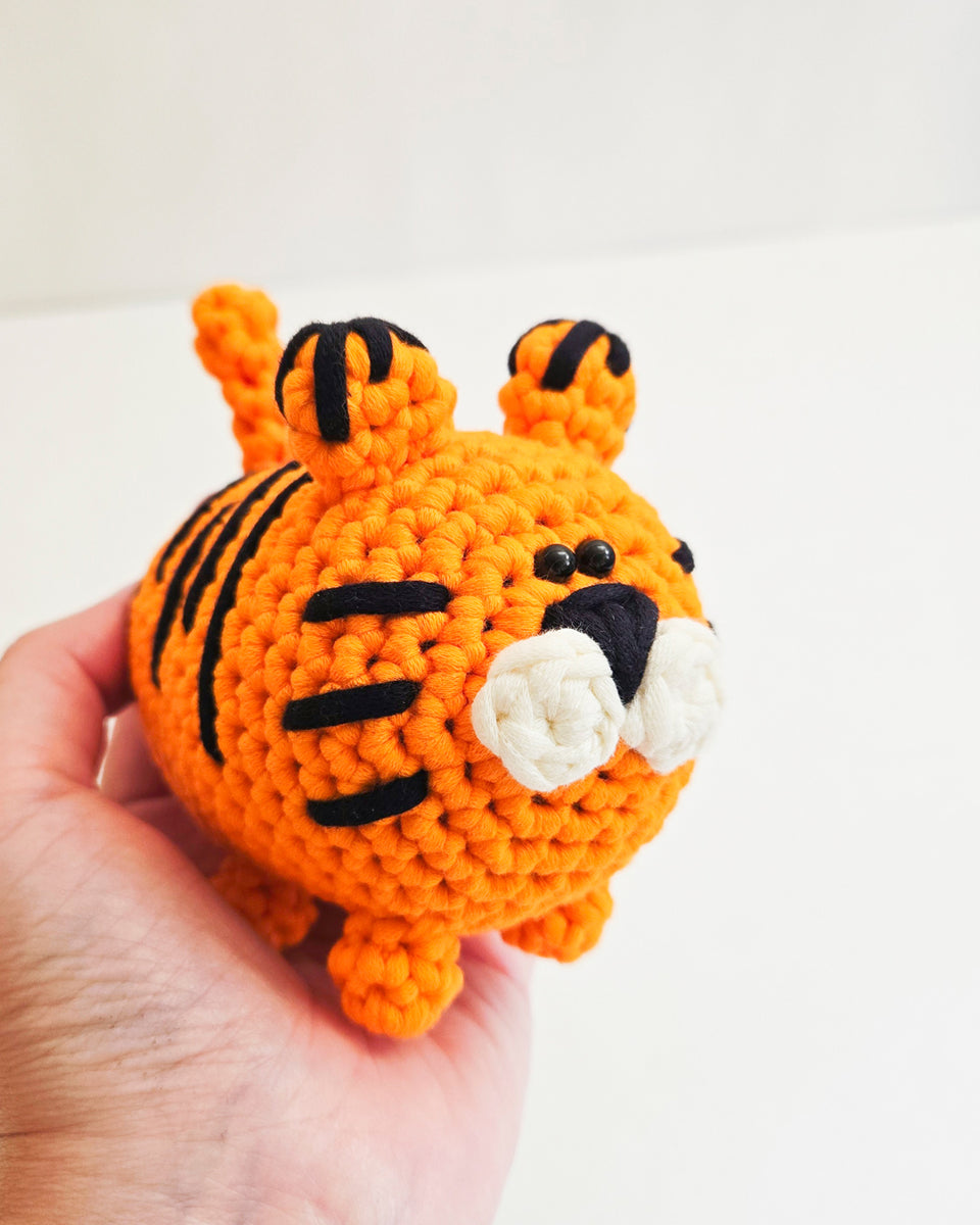 A small crocheted tiger toy being held in a person's hand against a plain light background.