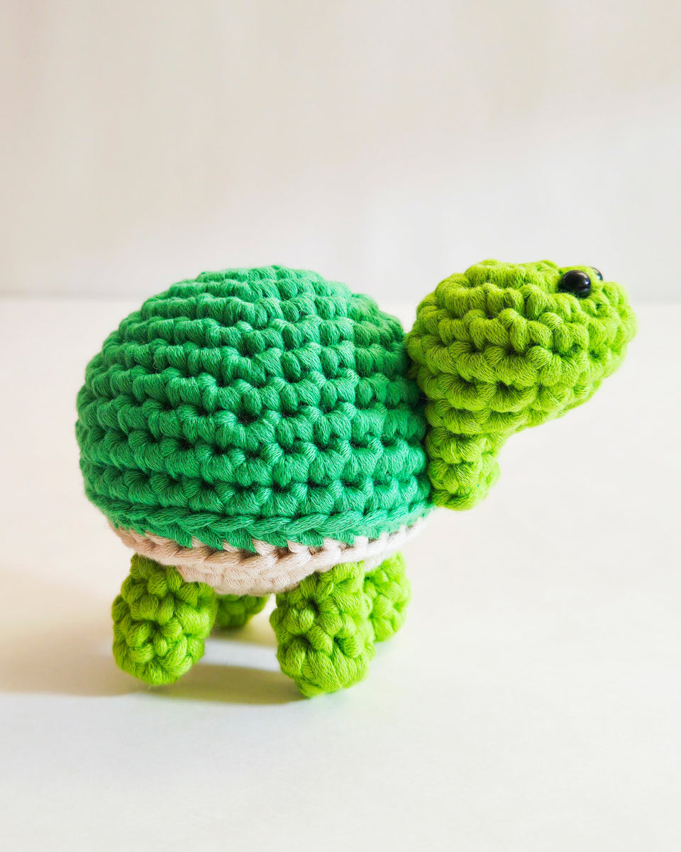 A handmade crocheted turtle toy made from a beginner crochet kit in shades of green with a beige underbelly, standing on a plain light background.