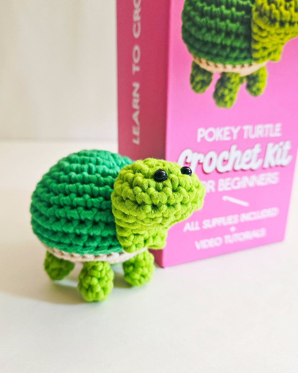 A small crocheted green turtle toy stands in front of a pink crochet kit box labeled 'Pokey Turtle Crochet Kit for Beginners' on a white surface.