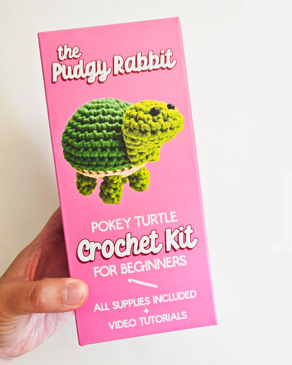 A hand holds a pink box for 'The Pudgy Rabbit Pokey Turtle Crochet Kit for Beginners' against a plain white background.