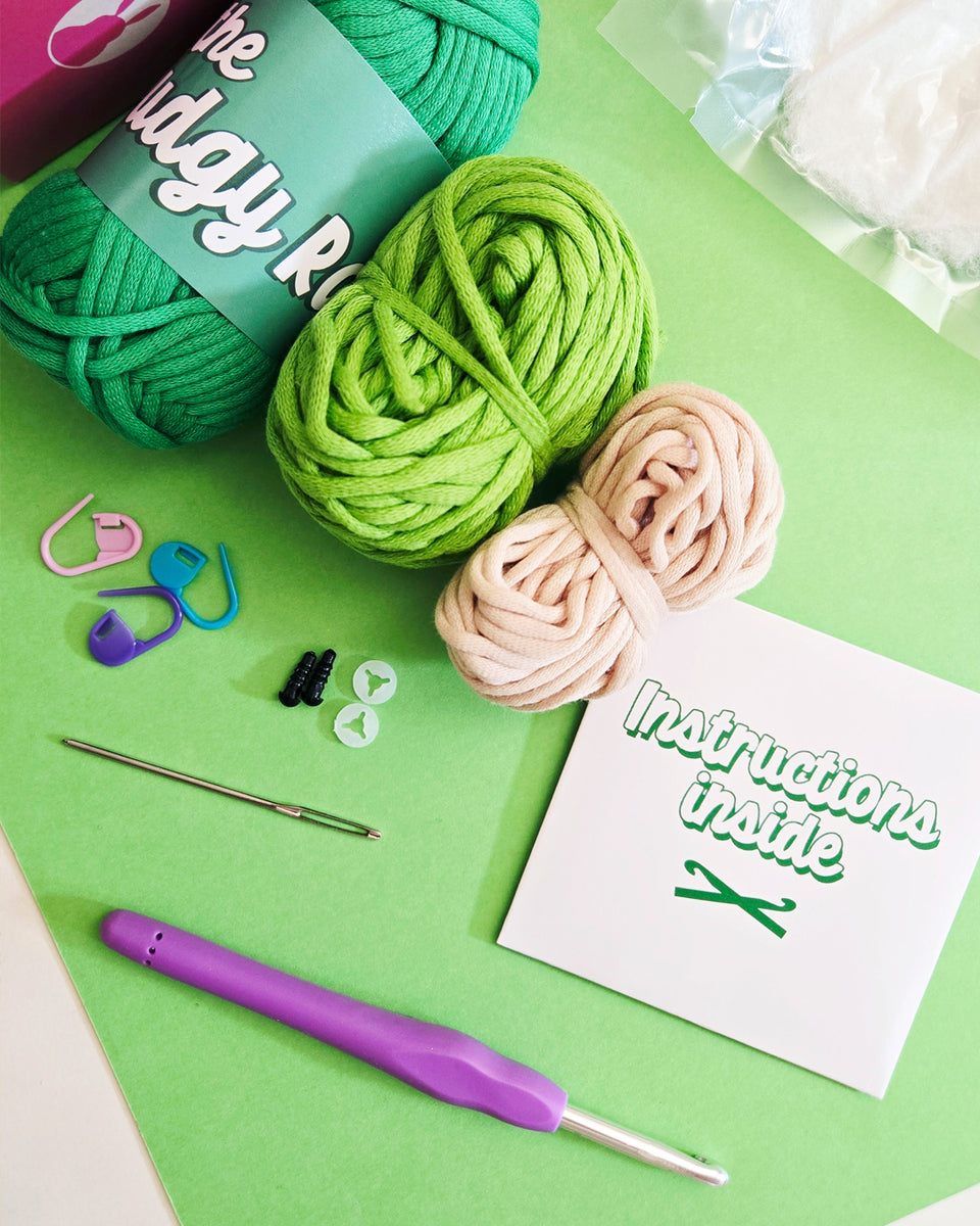 A flat-lay of a crochet kit on a bright green background, featuring three balls of yarn in green and tan, a purple-handled crochet hook, stitch markers, a metal yarn needle, and a card that reads 'Instructions Inside'.