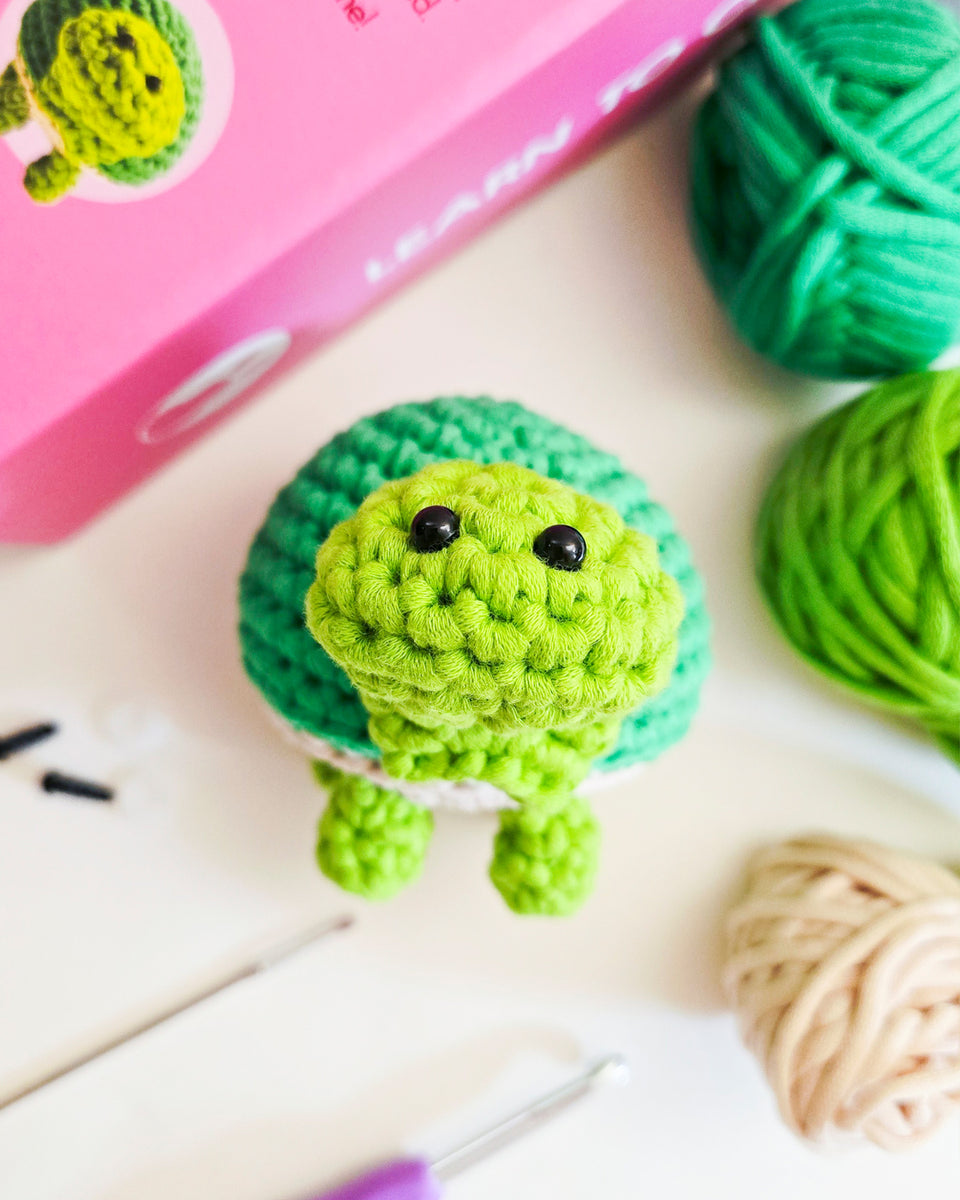 A crocheted green turtle toy with black bead eyes, styled on a white surface with yarn balls, a crochet hook, and a pink crochet kit in the background.
