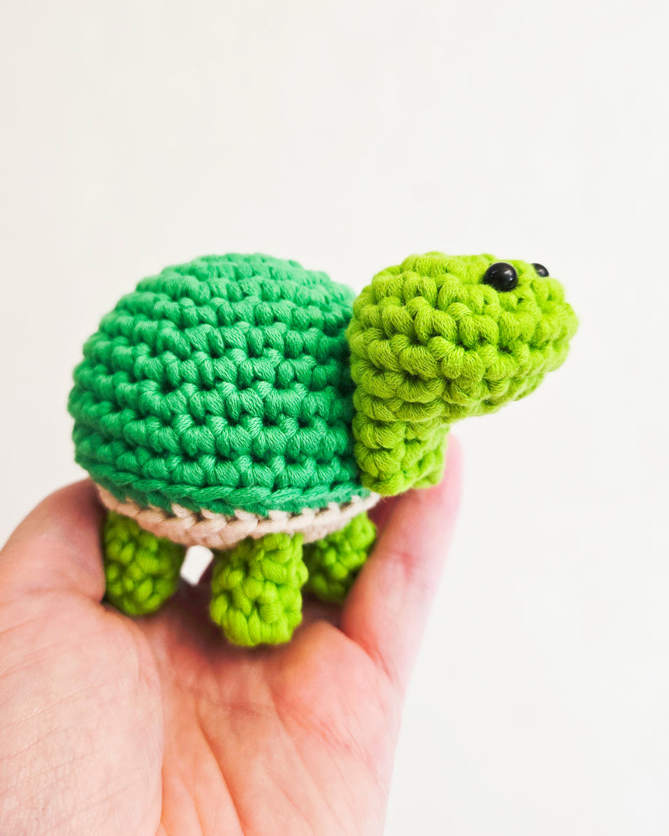 A small crocheted turtle toy held in a person's hand against a plain light background. The turtle has a bright green body and shell, a lime green head and legs, and a thin beige underbelly. Turtle crocheted from learn to crochet kit by The Pudgy Rabbit.