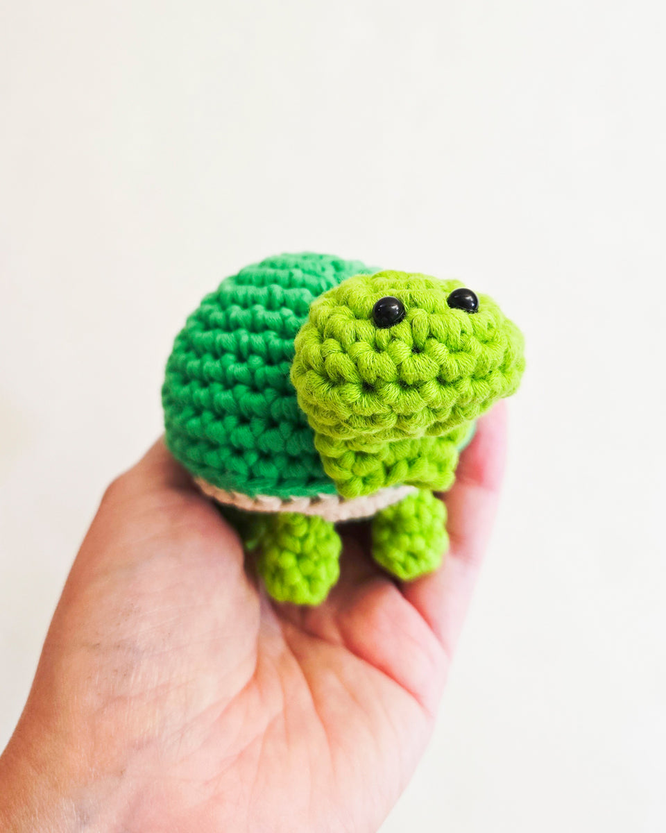 A small crocheted turtle toy held in a person's hand against a plain light background. The turtle has a bright green head and legs, a darker green shell, and small black bead eyes. Turtle made from a beginner's crochet kit by The Pudgy Rabbit.